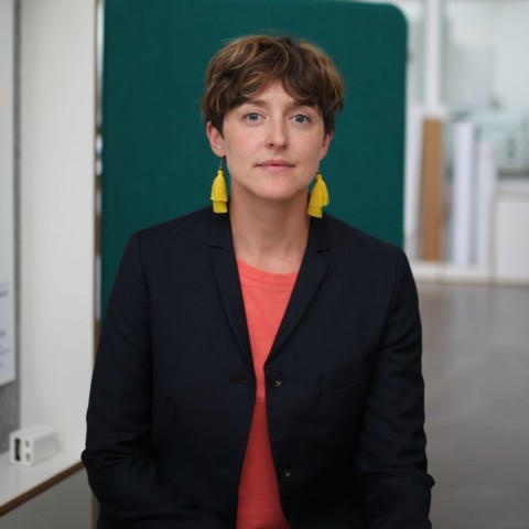 Elizabeth Calderón Lüning dressed in black suit and pink shirt with yellow earings and short hair in front of green background