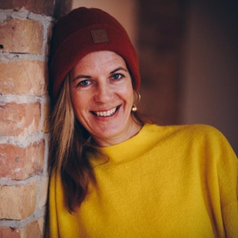 Katharina Naumann leaning against a wall with red hat and yellow pullover, smiling into the camera