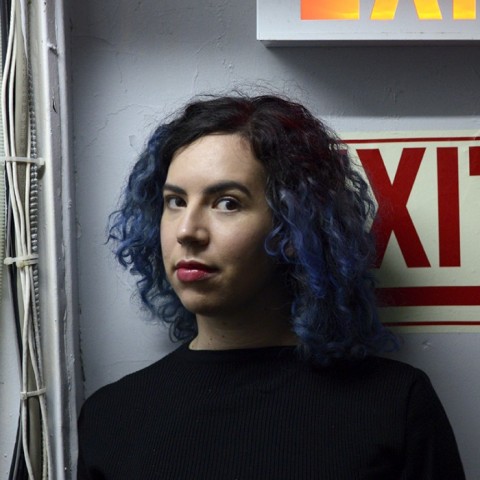 Caroline Sinders with curly blue and black hair in front of exit sign