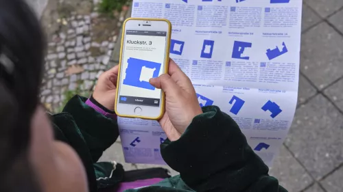 Ein Person hält ein Smartphone, auf dem Screen ist ein blauer Grundriss und eine Adresse.