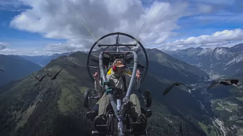 Mann in einem Leichtflugzeug über den Bergen, Vögel folgen ihm. 