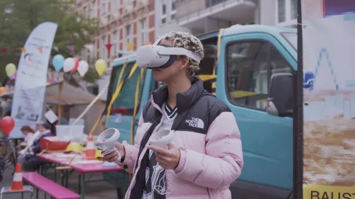 Eine junge Person hat ein VR-Headset auf und blickt zur Seite. In beiden Händen hält sie VR-Controller. Im Hintergrund ist ein Straßenfest mit Biertischen und einem VW-Bus zu sehen.