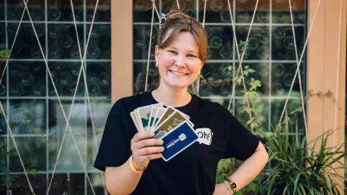Eine junge Frau in einem schwarzen T-Shirt steht mittig vor einer begrünten Glasfassade. In der rechten Hand hält sie ein Set bunter Karten. Sie lacht in die Kamera.