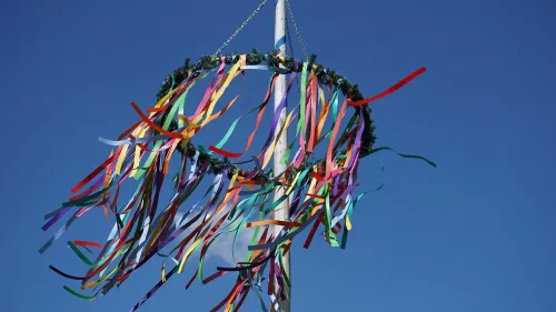 Das Bild zeigt einen traditionellen Maibaum mit bunten Bändern, die am oberen Ende des Maibaums befestigt sind. Die Bänder wehen im Wind und reichen in verschiedenen Farben wie Rot, Gelb, Blau, Grün und Lila. Der Maibaum selbst steht unter einem klaren, blauen Himmel und ist oben mit einem grünen Kranz verziert. 