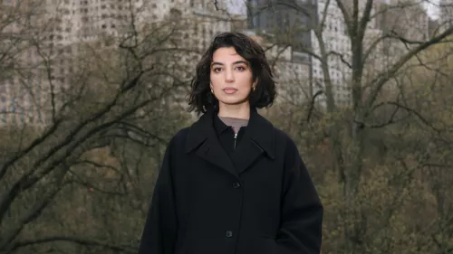Das Foto zeigt Aya Jaff in New York. Sie steht vor Bäumen und Gebäuden und trägt einen Mantel sowie kinnlanges Haar