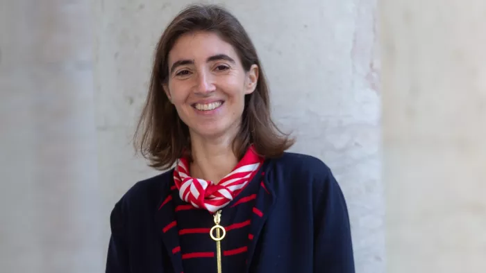 Chiara trägt schulterlange Haare, ein Halstuch und lächelt in die Kamera. Sie steht vor einer hellen Betonwand.