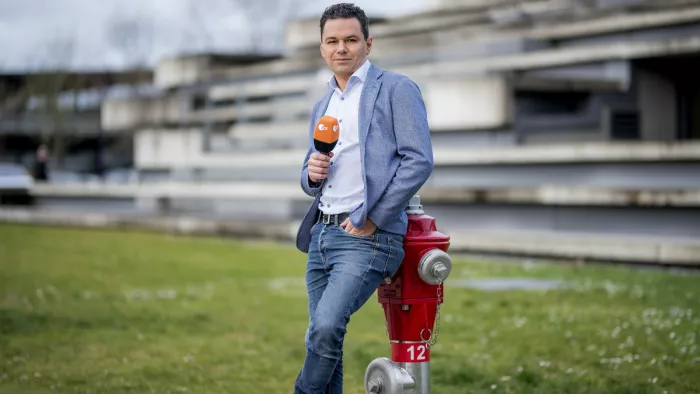Özden steht auf einer Wiese und lehnt sich an einen Hydranten. Er hält ein Mikrofon in der Hand.
