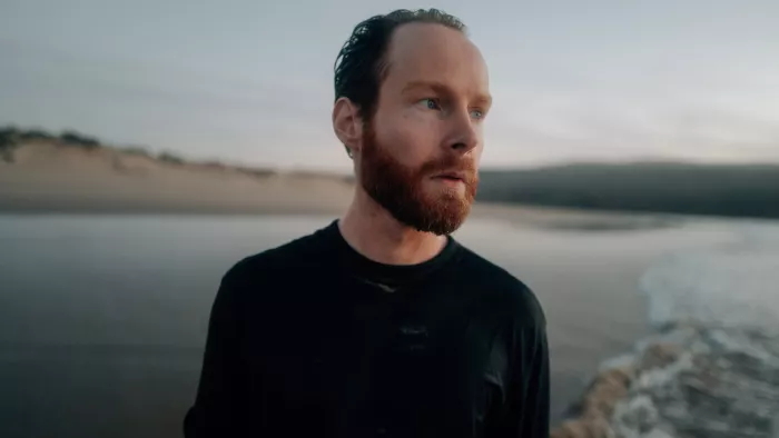 Enno Bunger trägt ein schwarzes T-Shirt und steht draußen in der Natur vor einem Fluss oder See.