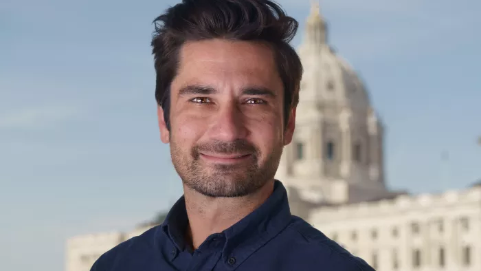 Casey Mock trägt kurze Haare und ein Hemd. Er schaut direkt in die Kamera. Der Hintergrund ist verschwommen und zeigt einen klaren Himmel und einem großen Gebäude. 