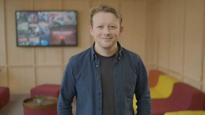 Philipp Lorenz-Spreen trägt kurze Haare, ein Hemd und darunter ein T-shirt. Im Hintergrund ist verschwommen eine Sofagarnitur und ein Bildschirm zu sehen. Er lächelt in die Kamera