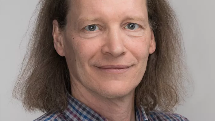 Portraitfoto von Christopher Coenen. Er trägt ein kariertes Hemd und posiert vor einem grauen Hintergrund. Seine Haare sind schulterlang