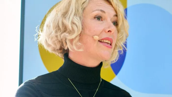 Das Foto zeigt Harriet Kingaby. Sie gestikuliert mit den Händen und trägt schulterlanges lockiges Haar so wie einen Rollkragenpullover und Mikrophon.