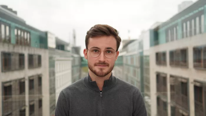 Portraitfoto von Maurice Höfgen. Er trägt kurze Haare, Bart, einen Pullover und Brille. Er schaut in die Kamera. Im Hintergrund sind auf beiden Seiten Gebäude zu sehen.