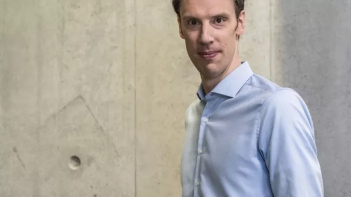 Foto von Björn Ommer, der ein Hemd trägt und in die Kamera schaut. Er steht vor einer Betonwand.