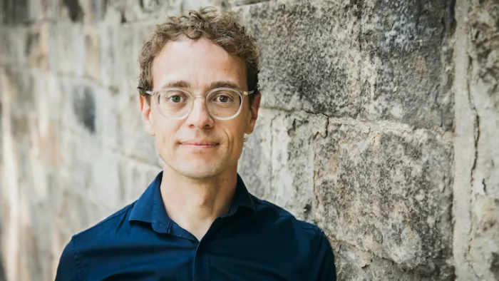 Foto von Simon Meier-Vieracker. Simon trägt kurze Haare, Hemd und Brille. Er steht vor einer Steinwand. 
