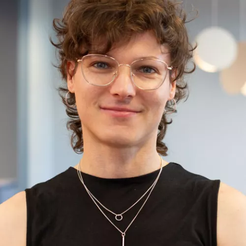 Portrait von Robyn, sie steht in einem schwarzen Oberteil vor einem unscharfen hellen Hintergrund und trägt eine Brille sowie eine zweireihige Kette.