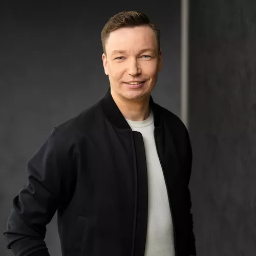 Porträt von Matthias Hölscher. Er lächelt und trägt eine schwarze Jacke über einem weißen T-Shirt