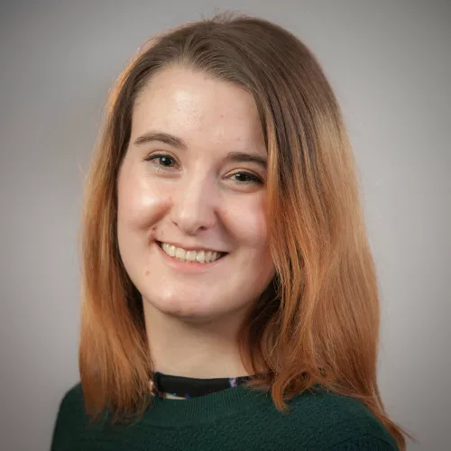 Das Bild zeigt eine lächelnde Frau um die 30, mit langen hellbraunen Haaren, heller Hautfarbe und braunen Augen.