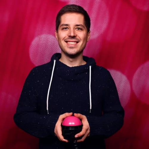 Robert Hecklau vor einer pinken Wand mit einem pinken Selbstauslöserbutton in der Hand. Er trägt einen blauen Wollpullover und lacht breit in die Kamera.