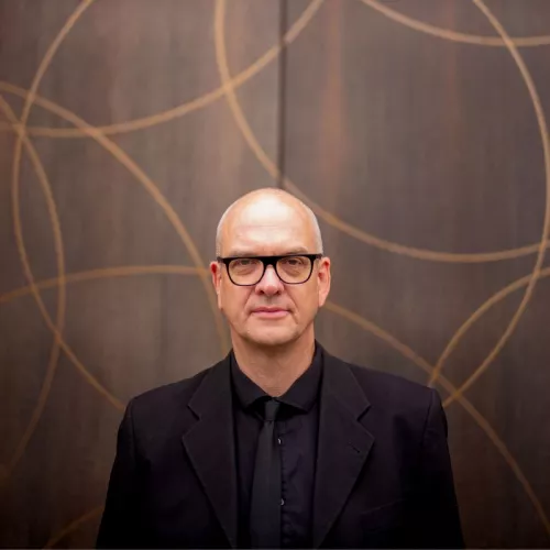 Boris Eldagsen ist in einem schwarzen Anzug gekleidet und trägt eine schwarze Brille. Er ist ein Mann mittleren Alters ohne Haare und steht vor einer braunen Tür mit goldenen Ornamenten.