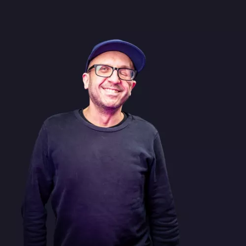 Johannes grinsend mit blauem Pullover, blauem BaseCap und schwarzer Nerdbrille.