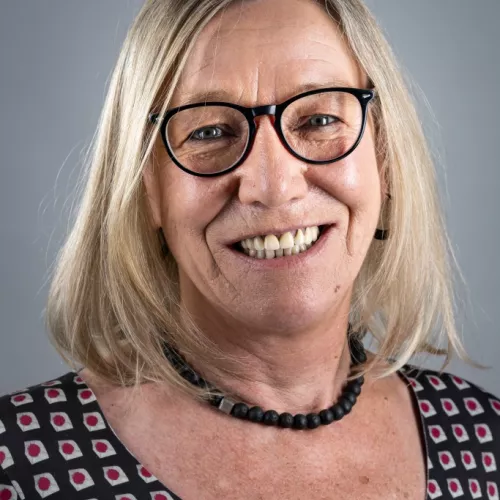 Das Foto zeigt eine lächelnde erwachsene Frau vor neutral grauem Hintergrund. Sie hat schulterlanges, hellblondes Haar. Sie trägt eine schwarze Brille mit runden, etwas dickeren Rahmen. Sie trägt ein gemustertes Oberteil mit quadratischen Formen in Schwarz, Weiß und Rot. Um den Hals hat sie eine dunkle Halskette aus matten, runden Perlen. 