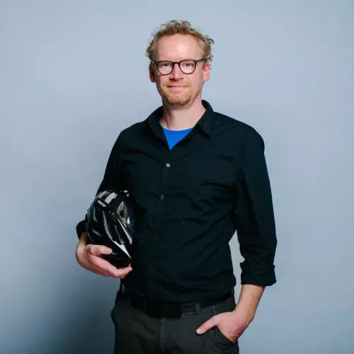 Portraitfoto Henning Krause: Ein weißer Mann Mitte vierzig mit Dreitagebart und Brille im schwarzen Oberhemd mit leicht hochgekrempelten Ärmel steht in der Mitte des Bilds und lächelt leicht in die Kamera. Unter dem rechten Arm hält er einen schwarzen Fahrradhelm.