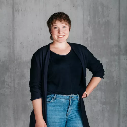 Eine Frau mit kurzen dunkelblonden Haaren steht vor einer Betonwand. Sie hat die linke Hand in die Hüfte gestützt und lacht in die Kamera. Sie trägt Blue Jeans und einen langen dunkelblauen Blazer.