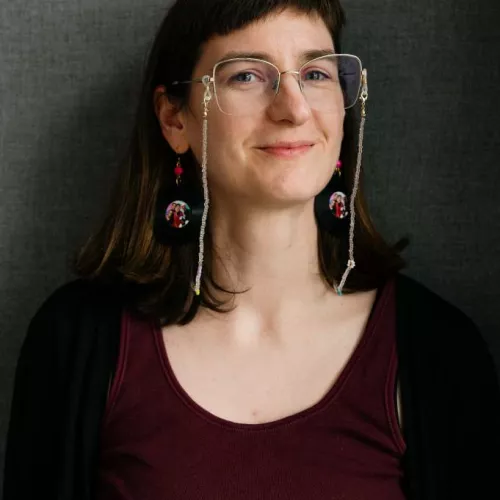 Paula schaut von rechts in die Kamera und trägt eine große Brille mit Brillenkette. 