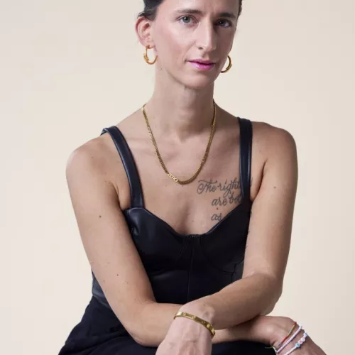 Auf dem Bild ist die Autorin Gazelle Vollhase zu sehen, sitzend auf einem Stuhl. Sie trägt ein ledernes Top mit Trägern, pinken Lippenstift, goldene Ohrringe und eine goldene Kette um den Hals. Auf der linken Brust hat sie einen Schriftzug tätowiert. 