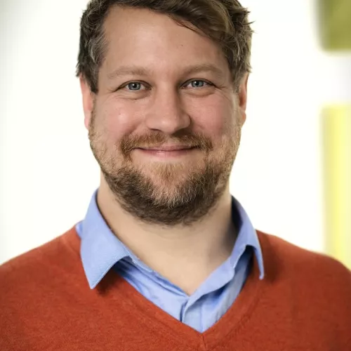 Portraitfoto des Speakers, roter Pullover, blaues Hemd, grüner und weißer Hintergrund.