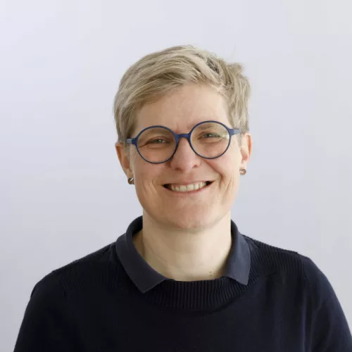 Porträt einer lächelnden Person mit kurzen blonden Haaren und runder blauer Brille, die vor einem hellen, neutralen Hintergrund in die Kamera schaut.
