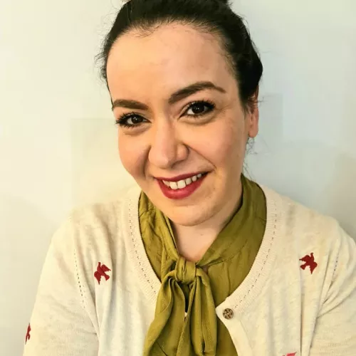 Frau mit hochgebundenem schwarzem Haar und roten Lippen in grüner Bluse mit großer Schleife und hellem Cardigan