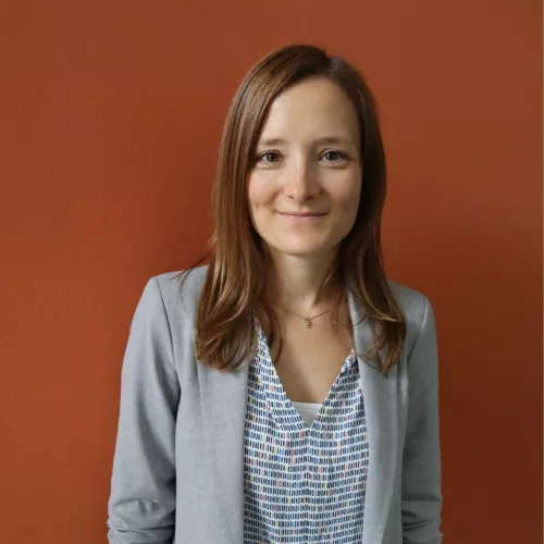 Carolin Rössler (Frau) mit braunen Haaren trägt einen grauen Blazer und steht vor einem braunen Hintergrund; sie lächelt in die Kamera