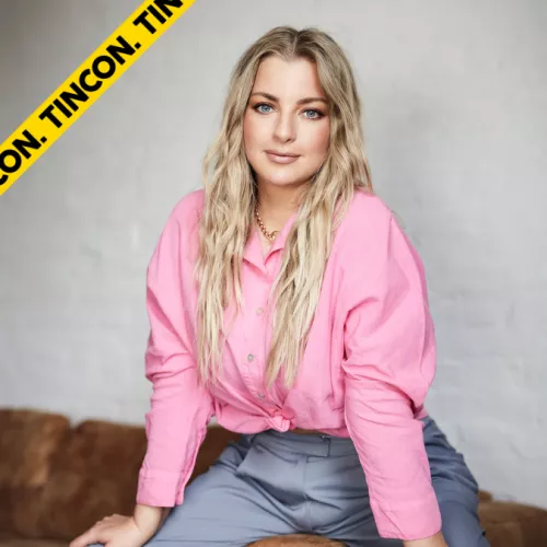 Laura sitzt auf einem Hocker. Sie hat lange, offene blonde Haare, trägt eine rosa Bluse und Jeans.