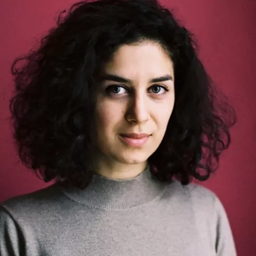 Portraitfoto einer PoC Frau mit schwarzen schulterlangen Locken und seitlichem Blick zur Kamera, leicht lächelnd.