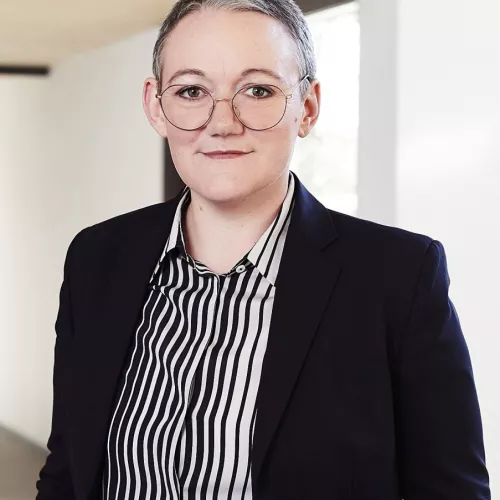 Judith Muster trägt einen schwarzen Anzug, ein schwarz-weißes Hemd, kurze Haare und eine Brille. 