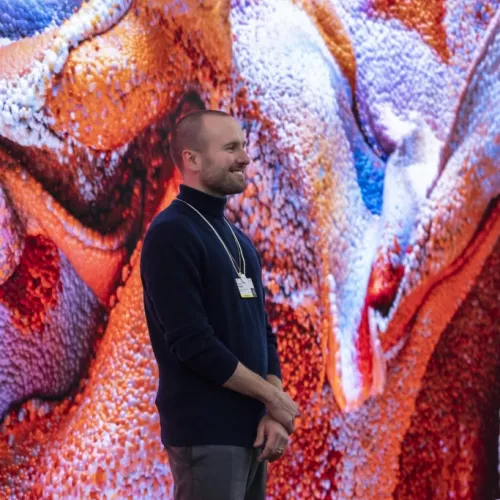 Florin Hasler in front of a colorful redish background (artwork Machine Hallucinations by Refik Anadol)
