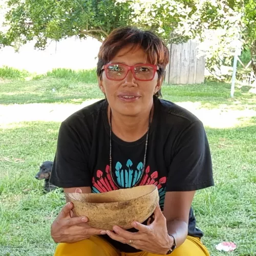 Ana Vilacy Galucio is sitting holding a gourd in both hands. She is wearing a black T-shirt with an image of an indigenous headdress. A dog is lying behind her. Green background of grass and trees behind.
