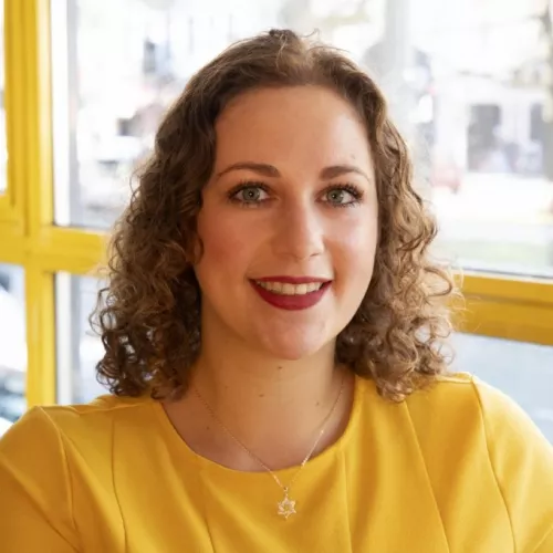 Dalia Grinfeld schaut direkt und freundlich in die Kamera. Sie hat schulterlanges lockiges Haar und trägt ein gelbes Oberteil. Sie sitzt vor einer Fensterfront.