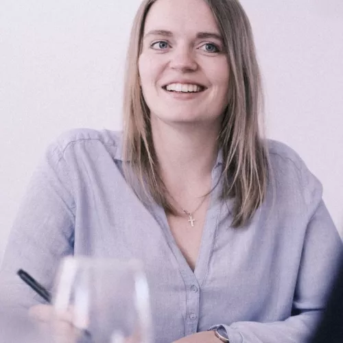 Das Bild zeigt eine lächelnde junge Frau mit langen, blonden Haaren und einer hellblauen Bluse, die an einem Tisch sitzt.