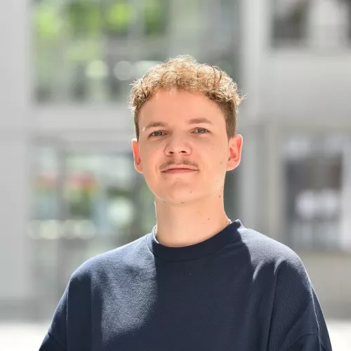 Junger Mann mit lockigen Haaren und leichtem Schnurrbart schaut freundlich in die Kamera