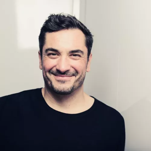 Porträt von Alexander Scheidt. Er schaut lächelnd in die Kamera und steht vor einem hellen Hintergrund. Er trägt einen schwarzen Pullover. Seine Haare sind braun und kurz geschnitten.