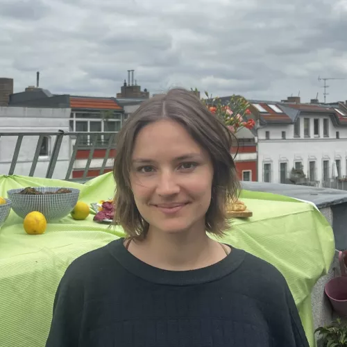 Das Bild zeigt eine junge Frau mit einem freundlichen Lächeln, die auf einer Terrasse steht, mit weiteren Dächern und Schornsteinen im Hintergrund.