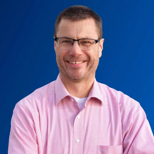 Andreas Maurer (Mann mit Brille, deunkelblonde Haare, trägt ein rosa Hemd, vor blauem Hintergrund)