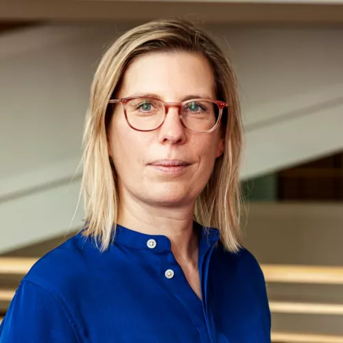 Antonia Götsch trägt eine blaue Bluse, blonde Haare und eine Brille. Sie schaut in die Kamera. 