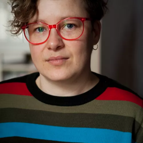 Porträit einer weiblich gelesenen Person mit kurzen Haaren, mit roter Brille und gestreiftem Pulli