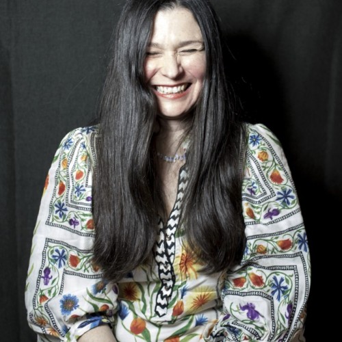 A picture of Belina Raffy being very smiley, and wearing a colourful silk shirt and jeans.
