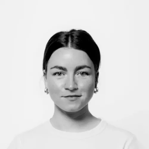 Schwarz-weiß Bild einer Frau, die direkt in die Kamera schaut. Sie hat braune Haare, trägt silberne Ohrringe und ein weißes T-Shirt.