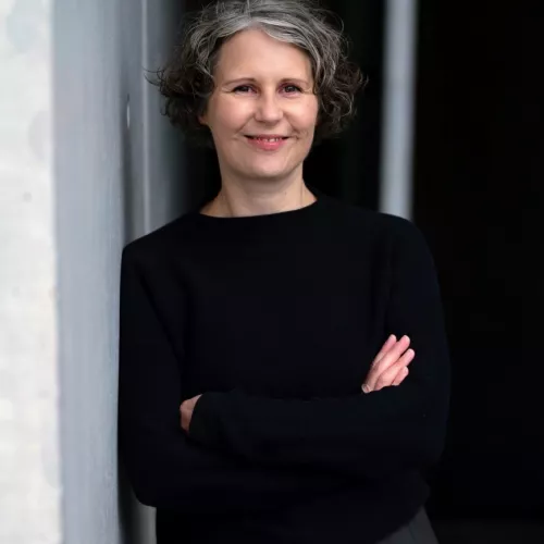 Birgit steht mit verschränkten Armen an eine Wand gelehnt. Sie lächelt.